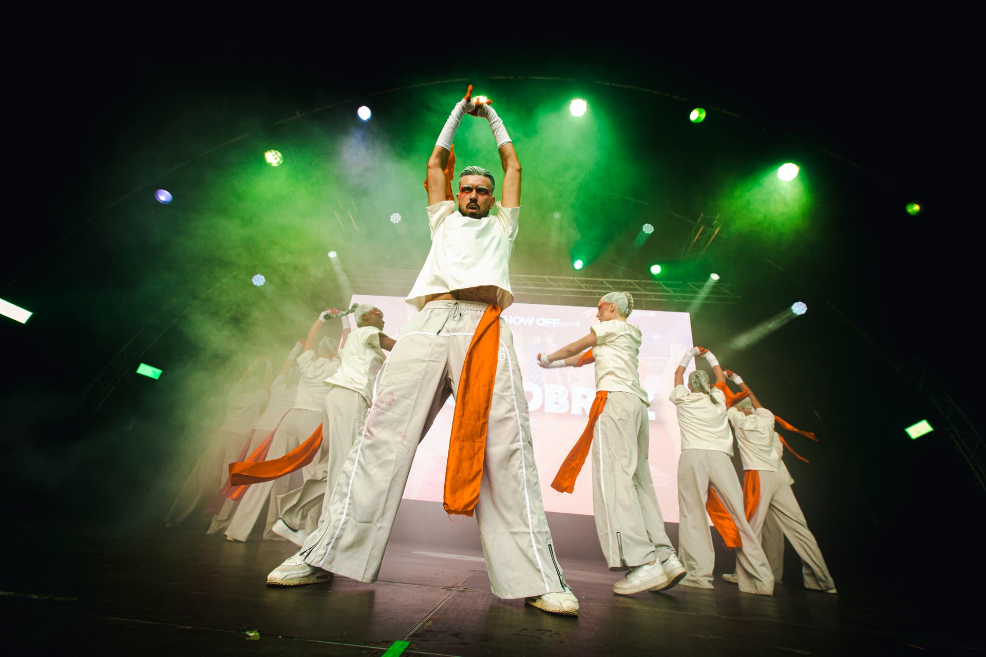 Dancers in flowing white outfits with orange sashes performing on The Show Off stage under green spotlights